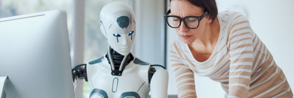 Woman in glasses studying a humanoid robot near a computer in a bright office setting