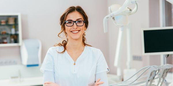 Smiling female dentist in white uniform standing in a modern dental clinic with equipment and monitor.