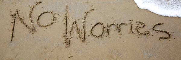 No worries written in sand near the ocean shore with a wave approaching the text.