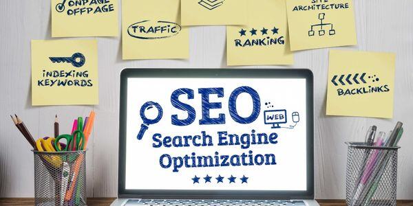 Laptop displaying SEO Search Engine Optimization surrounded by sticky notes with marketing keywords and office supplies.