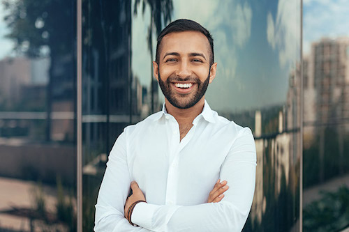 Confident man smiling with arms crossed wearing a white shirt outdoors in front of glass building