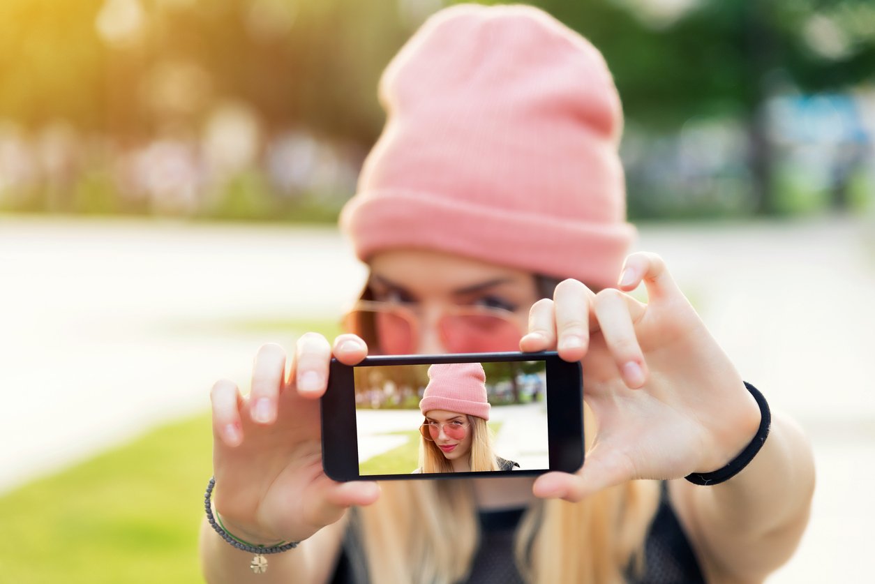 Young blonde in pink beanie taking a selfie for her Instagram Stories