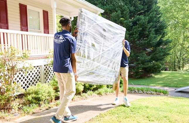 Two Midtown movers carrying a large wrapped item along a path outside a house with greenery surrounding the area. 