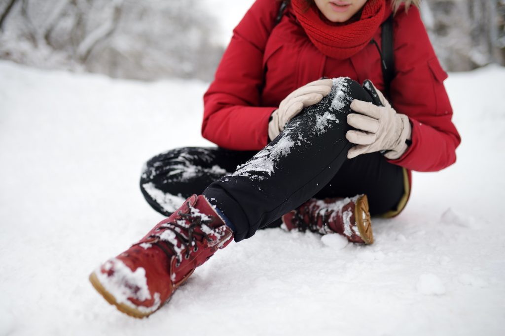 Shot of person during falling in snowy winter park. Woman slip on the icy path, fell, injury knee and sitting in the snow. Danger of season trauma.; blog: How to Avoid Common Winter Injuries